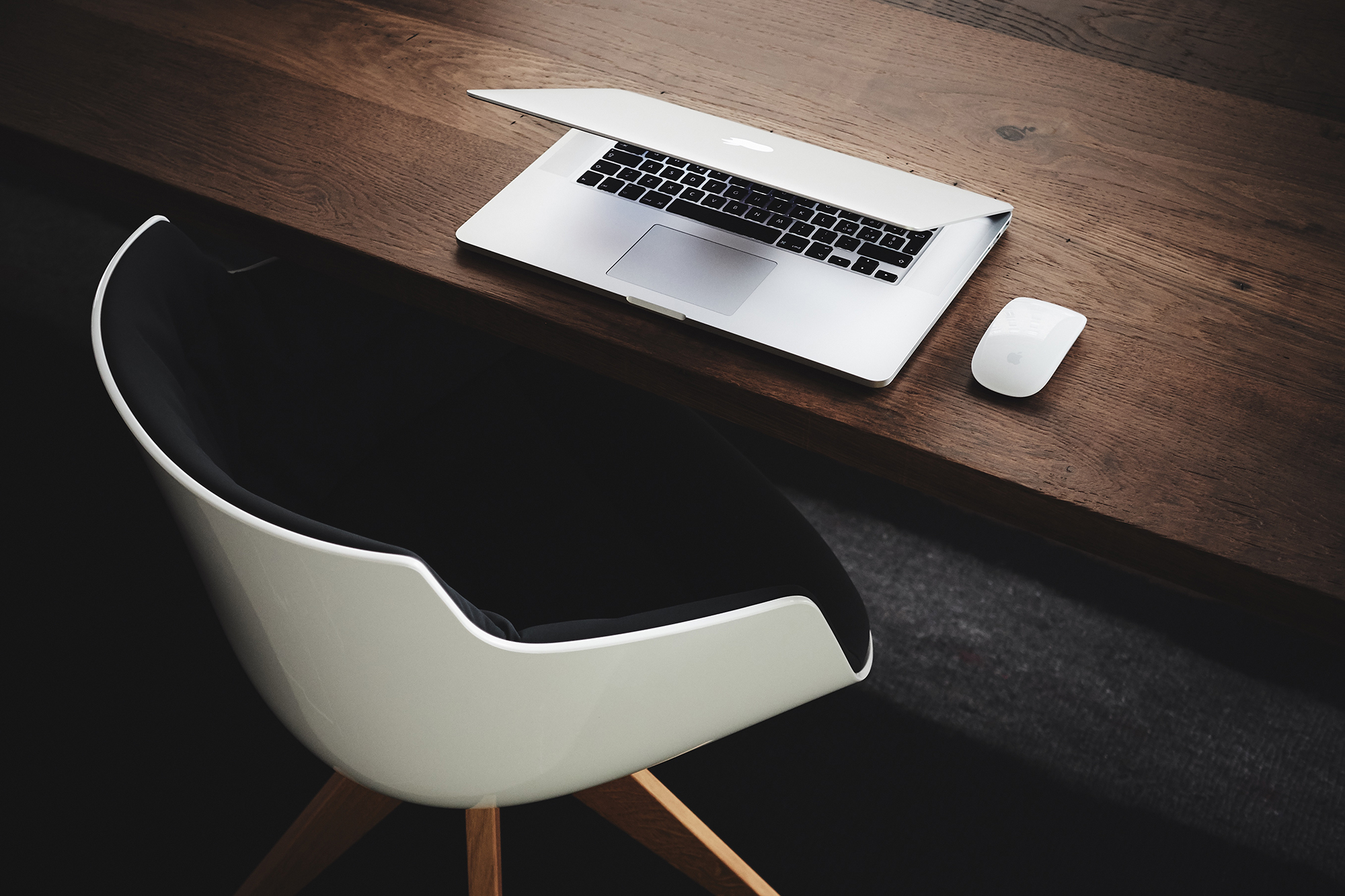 A Mac laptop with a Mac mouse on the table with a chair next to the table